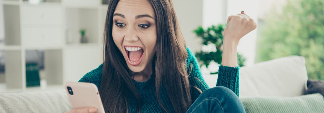 young woman sitting on her sofa playing on her mobile phone and very excited over a win