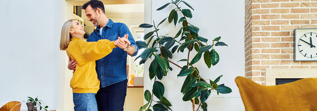 a couple smiling and dancing barefoot in their living room.  There is a large indoor potted plant in the room.