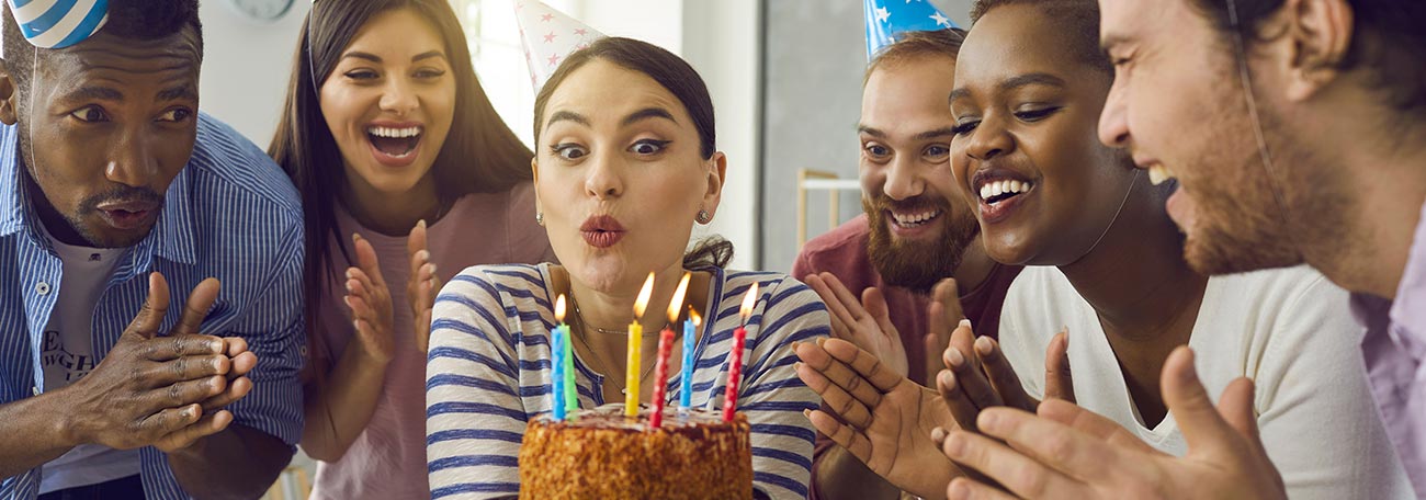 six twenty-something adults exuberantly celebrating a birthday as the birthday girl prepares to blow out the candles