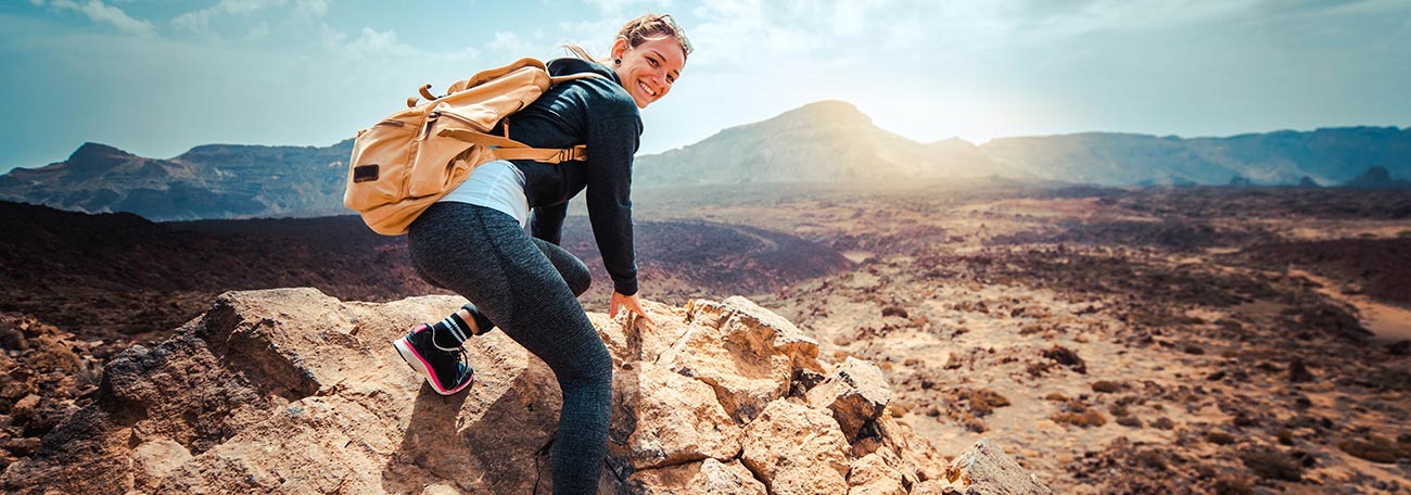 smiling attractive young woman with a backpack hiking in a mountainous area. In the background, the sun is reflecting off clouds