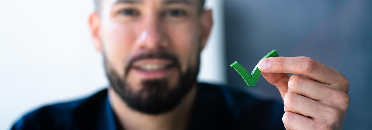 a smiling young man with a dark, well-cropped beard holding up a green check mark