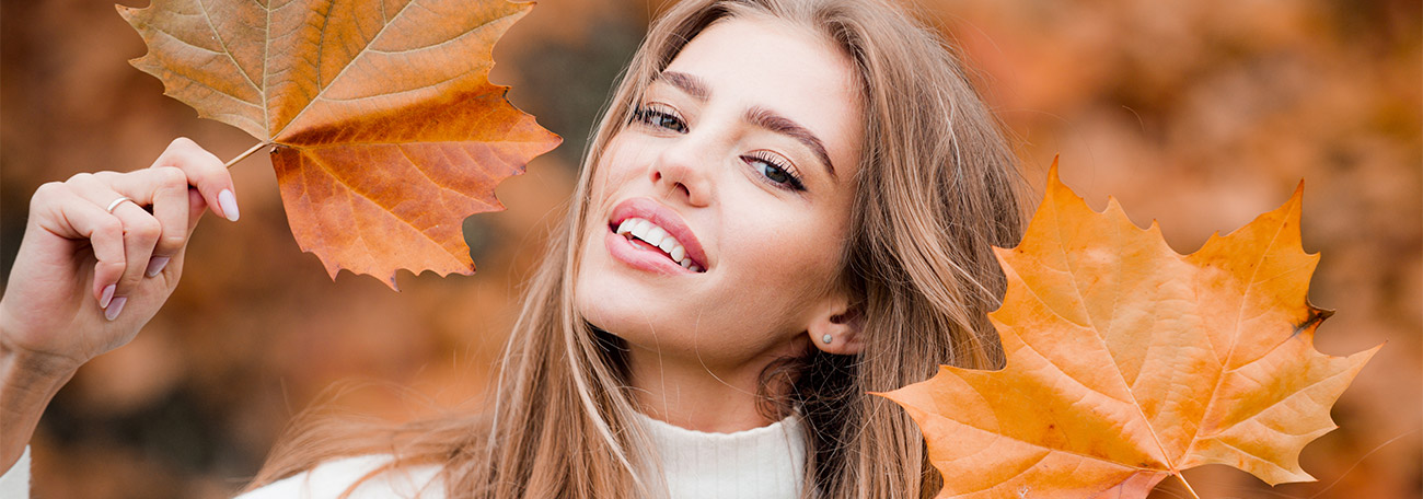 a young woman holding two large orange leaves from fall foliage and contrasting their color with her hair which is orange-red