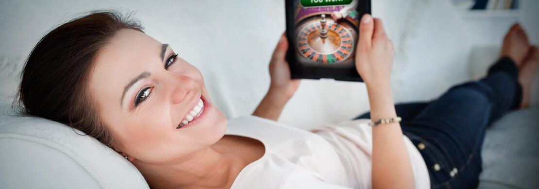 a smiling women relaxing comfortably on her sofa playing online casino games on her tablet